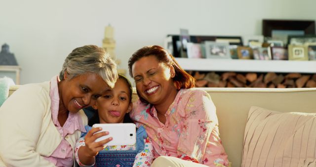 Depicts three generations of women, including a grandmother, mother, and child, joyfully taking a selfie while sitting on a couch at home. Ideal for use in family-themed projects, technology use in everyday life, or demonstrating multigenerational bonding and togetherness.