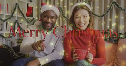 Merry Christmas couple wearing Santa hats smiling on cozy decorated couch holding mug