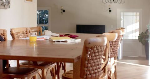 Sunlit wooden dining table showing scattered notebooks, headset and orange juice for work from home