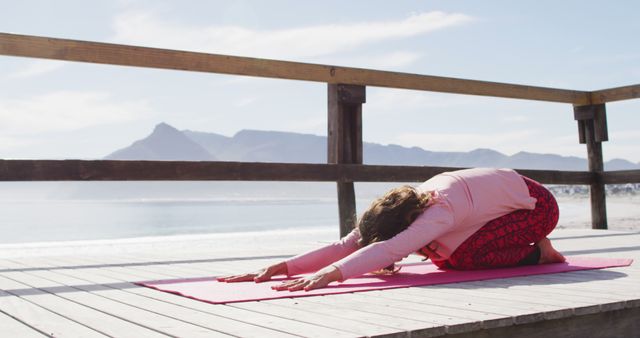 Woman practicing yoga on an outdoor wooden deck overlooking scenic mountains and ocean. She is in a pose on a yoga mat, surrounded by serene natural beauty, suggesting themes of health, relaxation, and tranquility. Ideal for wellness, exercise, travel, and lifestyle content.