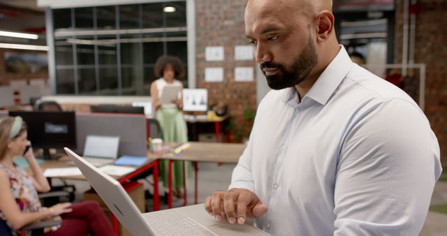 Man using laptop in a modern office while colleagues work in background. Suitable for themes on technology use in corporate environments, teamwork, and productivity in professional settings.