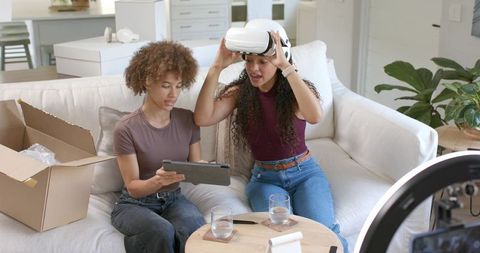 African American women testing VR headset and unboxing gadget on modern living room sofa