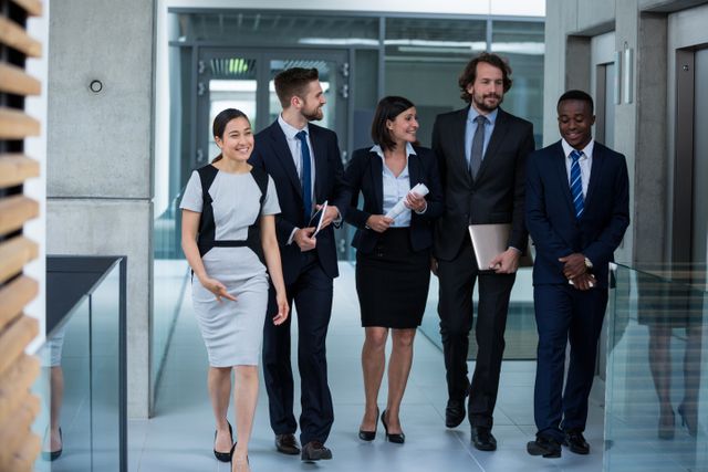 Diverse group of business professionals walking together in a modern office hallway, engaging in conversation. Ideal for use in corporate websites, business presentations, teamwork and collaboration themes, and professional development materials.