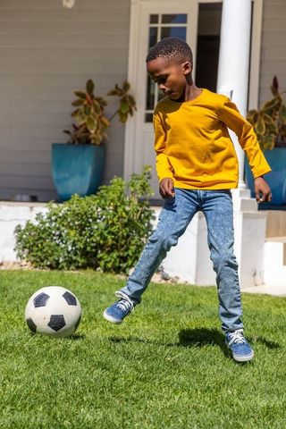 African american boy playing soccer on sunny suburban lawn wearing mustard shirt and jeans