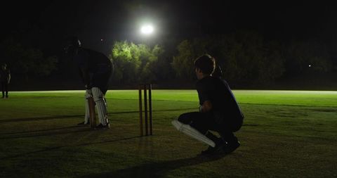 Wicketkeeper crouching behind stumps under floodlights watching batsman on night pitch