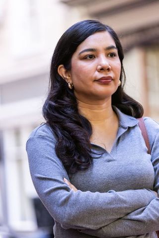 Pensive woman standing with arms crossed wearing gray knit polo and gold hoop earrings
