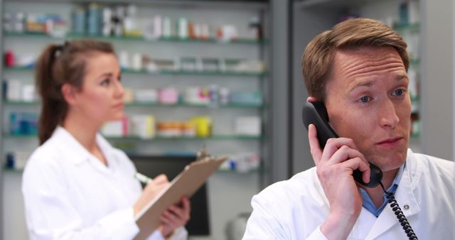 A Caucasian male pharmacist is on the phone, looking concerned, while a female pharmacist in the background checks inventory, with copy space. Their focused expressions and the pharmacy setting suggest they are dealing with an important matter related to patient care or medication management.
