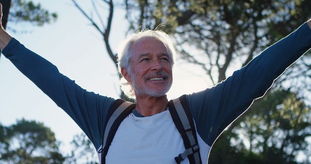 Senior man enjoying a hiking adventure in a forest, smiling and raising his arms joyfully. Ideal for advertisements, marketing materials, or content promoting active lifestyles, nature activities, and senior health and wellness.