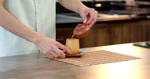 Man Pouring Tea from Teapot into Wooden Cup on Woven Placemats