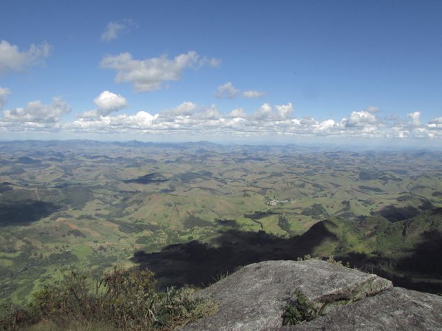 Panoramic view of sprawling green hills and valleys stretching into distance under a clear blue sky. White fluffy clouds dot sky, casting shadows on the terrain below. Suitable for travel brochures, nature and adventure websites, backgrounds for outdoor themes, and environmental projects focusing on natural beauty and preservation.