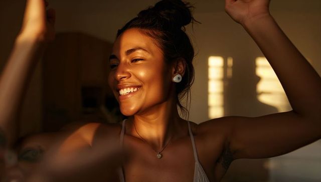 Smiling woman basking in warm golden light wearing earbuds, pendant and strappy top