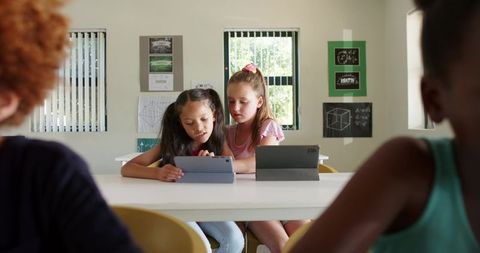 Schoolgirls collaborating on tablets in sunlit classroom learning math and technology