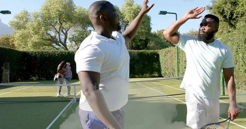 High-fiving tennis partners celebrating match win on sunny outdoor court, racket in hand