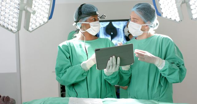 Surgeons in green scrubs and face masks discussing a surgical procedure while reviewing information on a tablet in an operating room. Bright overhead lights and medical equipment are visible. Ideal for healthcare, medical teamwork, technological advancements in medicine, and professional consultation imagery.