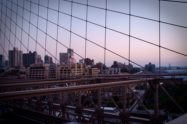 View of Manhattan's cityscape through the structural cables of Brooklyn Bridge during evening hours. Use for promoting travel destinations, showcasing urban architecture, or illustrating city life in New York.