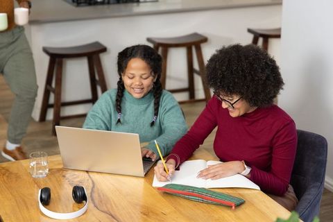 Diverse caregiver and child studying at kitchen table with laptop and notebook, home learning