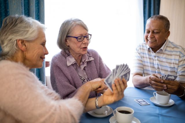 Seniors are enjoying a card game together at a table in a nursing home. They are smiling and appear to be having a good time. This image can be used for promoting senior living facilities, illustrating social activities for the elderly, or highlighting the importance of companionship and leisure in retirement.