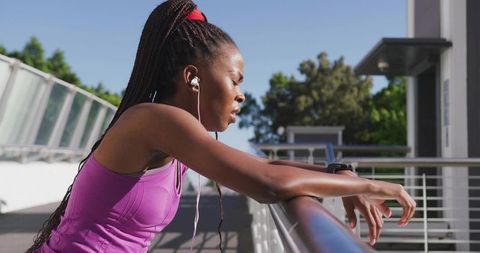 Woman catching breath leaning on railing after outdoor run wearing earbuds and pink tank