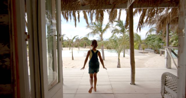 Image shows young girl walking barefoot towards tropical beach hut with thatched roof and palm trees in background, evoking feelings of vacation and adventure. Perfect for use in travel brochures, website banners for vacation rentals, ads promoting beach holidays, or social media posts about relaxing getaways.