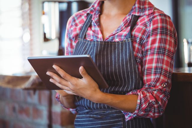 Waitress wearing plaid shirt and apron using a tablet in a cafe. Ideal for illustrating modern technology in the food service industry, digital order management, and efficient restaurant operations.