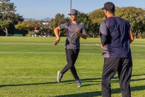 Cricket teammates practicing throws and catching drills on outfield in sunny session