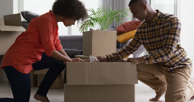 Young couple packing cardboard boxes together in a living room filled surrounded by other moving boxes and a couch with colorful cushions. Perfect for illustrating concepts related to relocation, settling into a new home, real estate transactions, and teamwork in household chores.