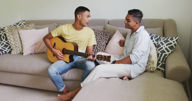 Smiling biracial gay male couple sitting on sofa one man playing guitar. staying at home in isolation during quarantine lockdown.