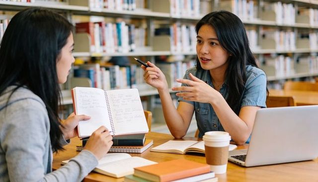 Two students collaborating and discussing notes at university library study table