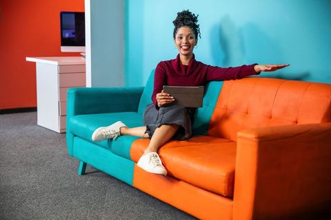 Smiling african american woman sitting on colorblock teal orange sofa using tablet
