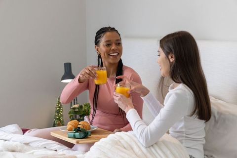 Multiracial friends toasting orange juice on cozy bed breakfast with gift and croissants