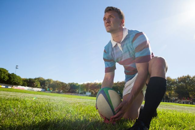 Rugby player kneeling on field getting ready to kick ball towards goal. Shows determination and focus under clear blue sky on a sunny day. Ideal for sports-related content, motivational themes, and athletic promotions.