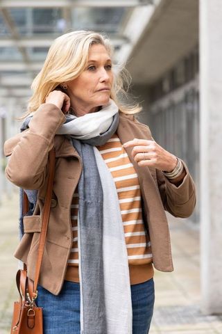 Middle-aged woman standing on urban walkway wearing striped sweater, scarf, camel coat carrying leat