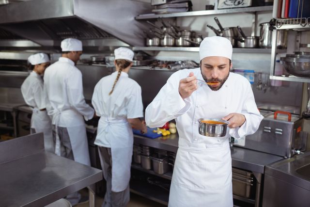 Chef tasting food from a pot in a professional restaurant kitchen while other kitchen staff are preparing ingredients in the background. Ideal for use in articles or advertisements related to culinary arts, restaurant management, professional cooking, and kitchen teamwork.