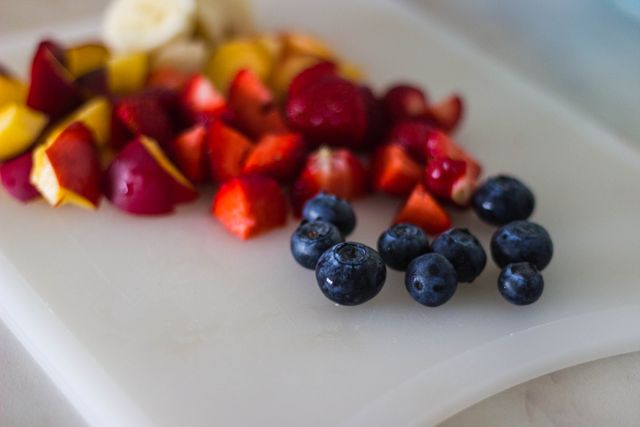 Freshly chopped fruits including blueberries, strawberries, and peaches are arranged on a white cutting board. Ideal for use in health food articles, diet recipe blogs, nutritional guides, and culinary websites. The variety of vibrant colors adds a visually appealing element to the image, making it suitable for advertisements and social media posts promoting healthy eating.