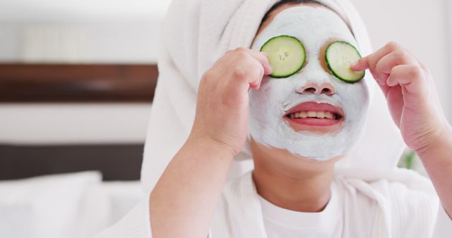 Child delighting in spa day experience with a face mask and cucumber slices over eyes. Great for themes involving self-care, relaxation, wellness, and family activities. Ideal for spa promotions, skincare advertisements, and lifestyle blogs.