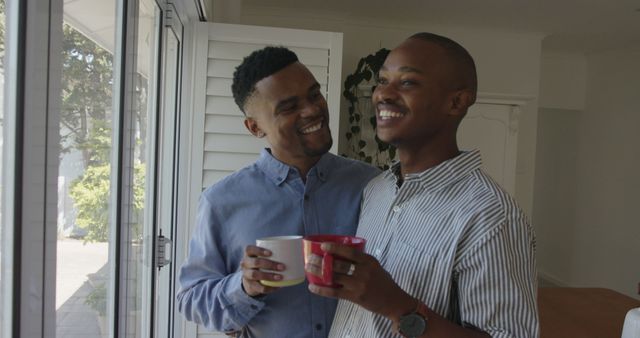 Happy african american gay male couple drinking coffee and laughing at home. Relationship, domestic life and togetherness, unaltered.