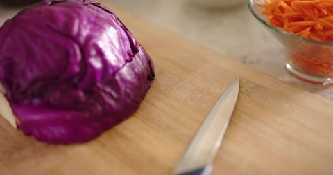Halved purple cabbage on wooden cutting board with knife and shredded carrot bowl