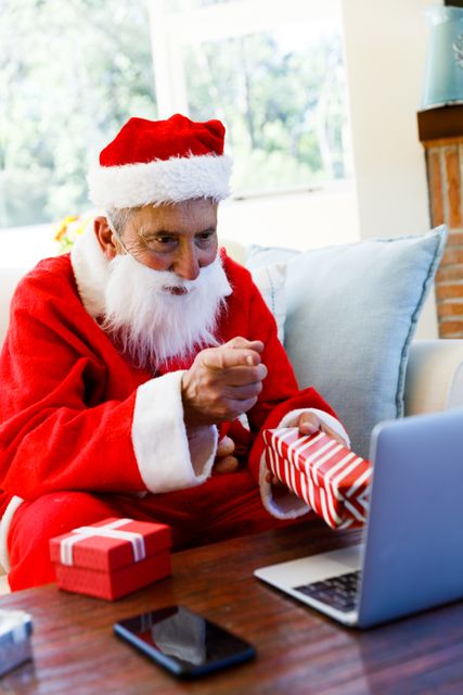 Side view of an old caucasian man wearing a santa clause costume holding out a christmas gift and pointing towards the laptop while on a video call. he's sitting on a couch in the living room