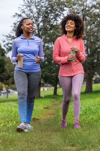 Two african american women walking and chatting in park holding insulated water bottles
