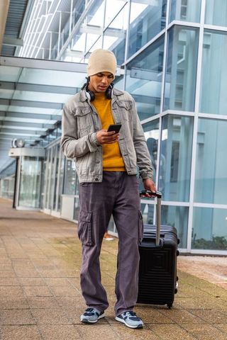 Urban traveler checking smartphone while pulling rolling suitcase at modern glass terminal