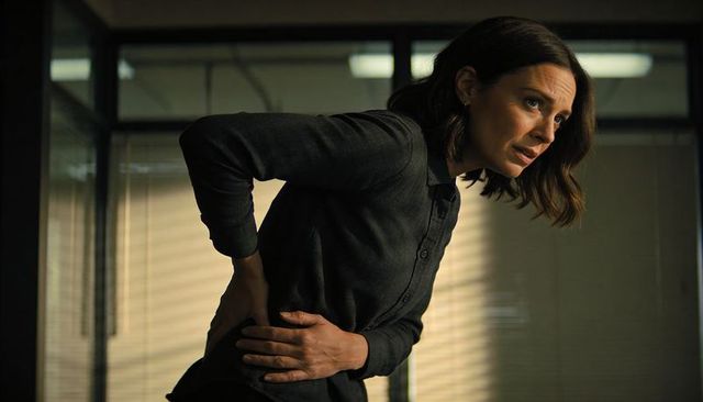 Professional woman clutching lower back in dim office, leaning forward from back pain