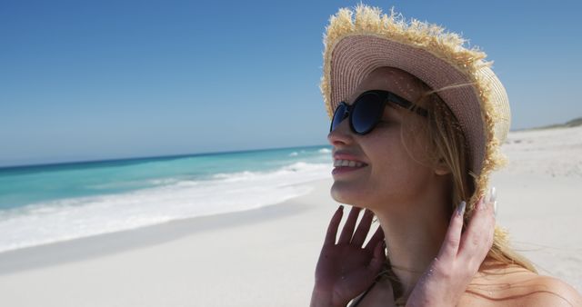 Adult woman smiling while enjoying a sunny day at the beach. Wearing a wide-brimmed hat and sunglasses, she appears to be in a relaxed and happy state. The ocean and sandy shore in the background suggest a perfect summer vacation. Ideal for websites, travel promotions, vacation packages, and lifestyle blogs focused on leisure and beach getaways.