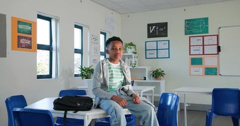 African teenage student wearing striped tee sitting on classroom table with backpack