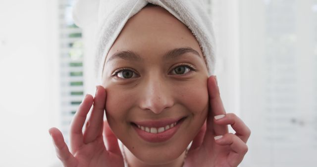 A close-up of a smiling woman with a towel wrapped around her head, touching her face with both hands. Perfect for promoting skincare products, beauty routines, spas, fresh and natural beauty, morning routine inspirations, or wellness and self-care articles.