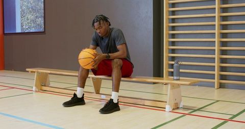 Young black man sitting on bench holding basketball and wearing wristwatch in gymnasium