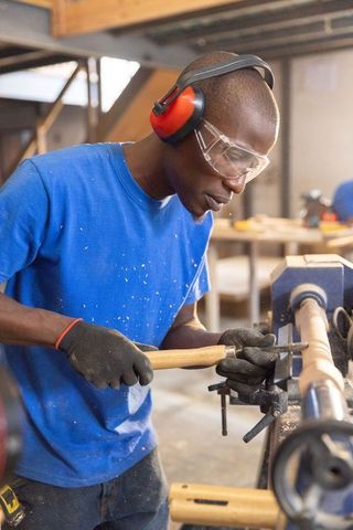 African american craftsman turning wooden spindle on lathe wearing safety gear