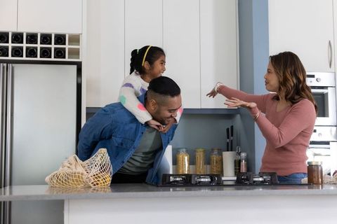 Playful family in modern kitchen, father carrying daughter piggyback while mother chatting