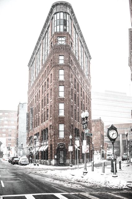 Vintage red brick building sits at street corner in snow-covered city. Snow blankets area, buildings in background. Captures essence of urban winter. Ideal for use in real estate promotions, historic district features, or cityscape presentations.