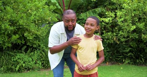 African american father encouraging smiling adolescent son in yard with tropical foliage