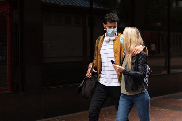 Young Caucasian couple walking in the city during the day, wearing face masks to protect against air pollution and COVID-19. They are embracing and talking, with the woman holding a smartphone. This image can be used to depict urban life during the pandemic, health and safety measures, or young adult interactions in public spaces.